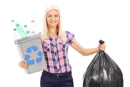 A young woman with long blonde hair, smiling and wearing a pink and blue checkered shirt, holding a grey recycling bin filled with various recyclable plastic bottles and containers in her left hand, while her right hand holds a large black rubbish bag. The background is plain white, emphasizing her and the waste collection items. The recycling bin features a blue recycling symbol, and the scene suggests an organised approach to waste disposal, relevant to general rubbish collection and recycling services offered by Commercial Waste Removal Hainault. The grey bin is made of sturdy plastic with a textured surface, and the plastic bottles inside are transparent or colored in green, blue, and clear finishes, with some capped and others open. The black refuse bag appears to be full and tied at the top, indicating it is ready for collection or disposal. The image conveys a professional and approachable attitude towards waste management, suitable for a rubbish removal service operating locally in Hainault or surrounding areas. The lighting is bright and even, highlighting the details of the waste containers and the woman's cheerful expression.