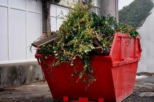 A large red skip bin filled with green waste, including branches, leaves, and other garden debris, situated on a paved surface near a white fence and concrete wall. The debris overflows from the top of the bin, with some branches and foliage leaning outward. The surrounding area is an outdoor space, possibly a driveway, with a partially visible building wall in the background. The scene is evenly lit with natural daylight, and the overall context suggests a waste removal task related to garden clearance, consistent with services provided by Commercial Waste Removal Hainault serving the local area.