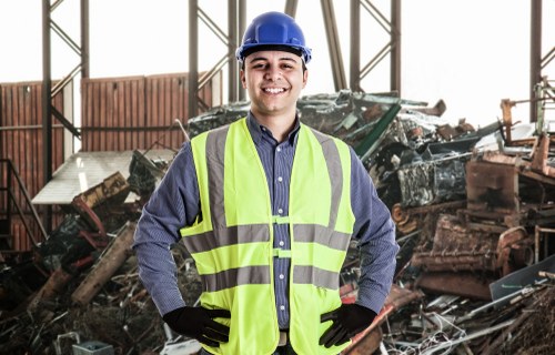 A young male worker at an indoor waste recycling or scrapyard facility, wearing a blue safety helmet and a high-visibility yellow vest over a blue long-sleeved shirt. He is smiling and standing with hands on hips in front of a large pile of mixed scrap metal, including car parts, sheet metal, and other metallic debris. The background features metal framework and partial structures of the warehouse or industrial building, with natural light illuminating the scene. The environment suggests a professional setting for rubbish or waste removal services, typical of operations in the Hainault area. The worker's attire and the visible waste materials represent a typical scene associated with commercial waste removal companies, such as Commercial Waste Removal Hainault, servicing local industrial and commercial clients.