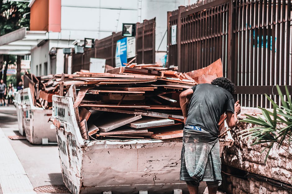 A man with dark, curly hair, dressed in a black T-shirt and grey plaid shorts, stands next to a large, open-top metal skip filled with a collection of reddish-brown roof tiles, wooden planks, and asso