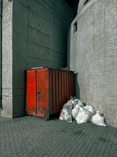 A man with dark, curly hair, dressed in a black T-shirt and grey plaid shorts, stands next to a large, open-top metal skip filled with a collection of reddish-brown roof tiles, wooden planks, and asso